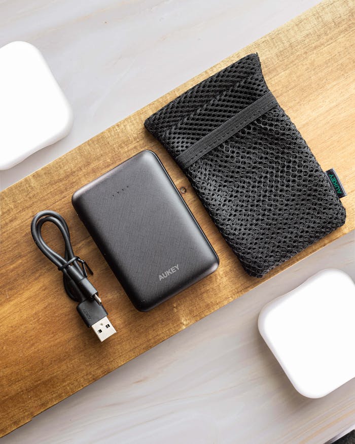 Flat lay of a black power bank with a USB cable and mesh pouch on wood.